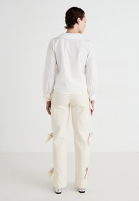 Camisa blanca de cuello con mangas abullonadas, combinada con pantalones crema que presentan acentos de lazada en el tobillo. Tejido texturizado y corte a medida.