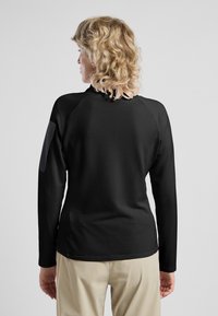 Zwarte languit-top met geribbelde textuur en raglanmouwen, met een gladde grijze patch op de bovenste linkerarm.