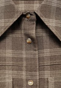 Chemise à carreaux marron avec col boutonné, deux boutons, et tissu texturé. Des nuances claires et foncées composent le motif à carreaux.