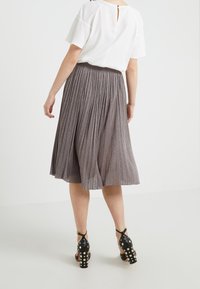 Gonna midi plissettata in un tessuto metallico grigio lucido, abbinata a una blusa bianca. Tacchi alti neri con dettagli borchiati. Vista posteriore.