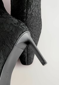 Černá vysoká botka na podpatku s látkou s krajkovým vzorem, špičatou špičkou a elegantním, lesklým černým stiletto podpatkem.