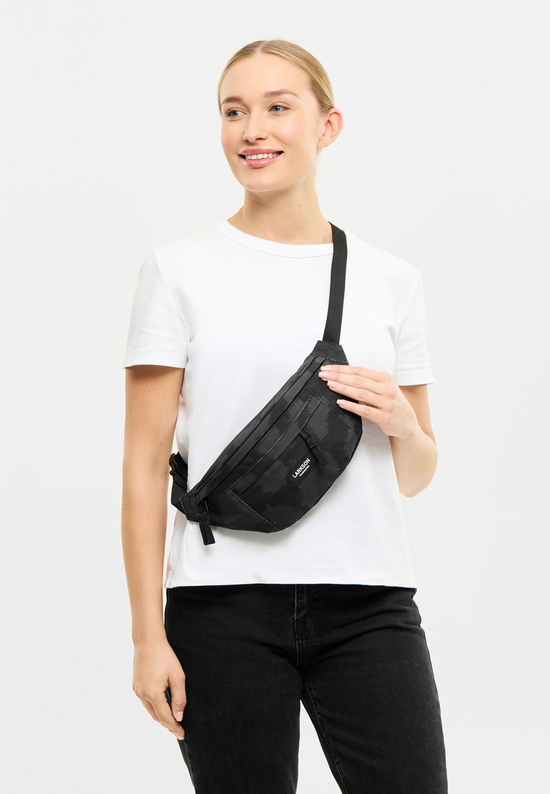 Borsa a fanny pack nera realizzata in tessuto testurizzato, dotata di una tasca frontale con zip, spallaccio regolabile e un logo sulla parte anteriore.