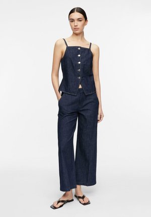 Kobieta stojąca z rękami w kieszeniach, ubrana w ciemny denimowy bezrękawnik zapinany na guziki, szerokie spodnie i czarne sandały z kwadratowym noskiem.