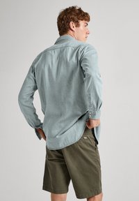 Camicia in denim blu chiaro con maniche lunghe, polsini con bottoni e orlo curvo, abbinata a pantaloni corti verde oliva con tasche laterali.