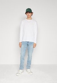 Vit långärmad skjorta med gult tryck, ljusblå jeans och vita sneakers med svarta accenter. En grön keps fulländar outfiten.