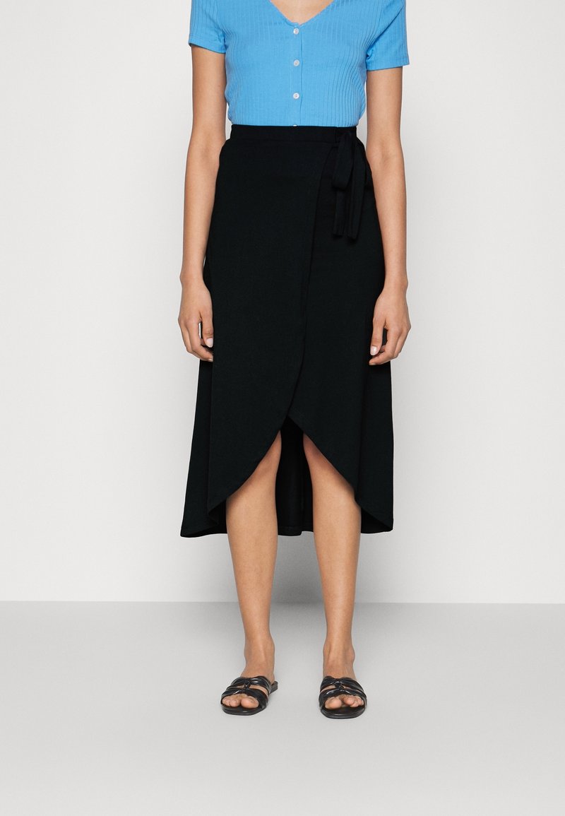 Femme vêtue d'un haut bleu à manches courtes avec boutons, d'une jupe portefeuille noire avec lien, et de sandales noires ouvertes à l'avant, debout devant un fond uni.