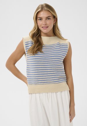 Młoda kobieta o długich blond włosach, ubrana w bezrękawnik z beżowego dzianego materiału z niebieskimi paskami oraz białe, luźne spodnie, uśmiecha się, mając rękę na biodrze.