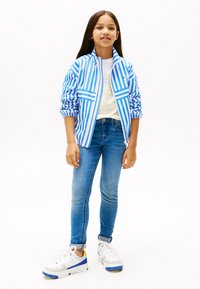 Chaqueta azul claro y blanca a rayas con cremallera sobre una camisa amarilla a rayas, combinada con jeans azules y zapatillas blancas con acentos azules y grises.