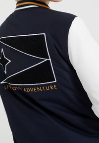 Marinblå varsityjacka med vita ärmar, prydd med en strukturerad svart geometrisk patch och broderade "LEVA DITT ÄVENTYR" i guld.