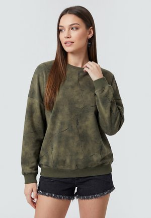 Ung kvinde med langt brunt hår iført grøn marmoreret sweatshirt og sorte frynsete denimshorts, kigger til venstre.