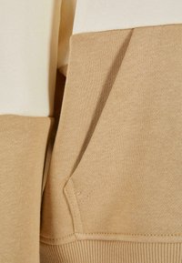 Felpa color block beige e crema che presenta una tasca frontale, orlo a costine e una texture liscia. Dettagli di cucitura visibili in tutto il capo.
