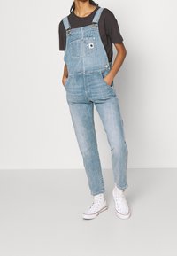 Lichte blauwe denim overall met verstelbare banden, een voorzak en taps toelopende pijpen, gecombineerd met een zwart t-shirt en witte sneakers.