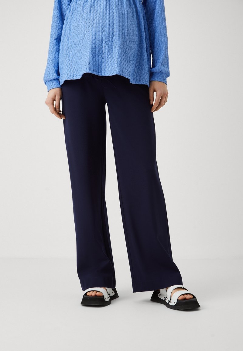 Personne portant un pantalon large bleu marine, un haut à manches longues bleu texturé et des sandales noires et blanches ouvertes à l'avant, debout sur un sol blanc.
