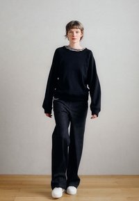 Melnraudzējs sporta krekls ar apaļu kakla izgriezumu, kopā ar tumšiem džinsa džinsiem. Modele valkā baltus sporta apavus. Vienkāršs, brīvs dizains.