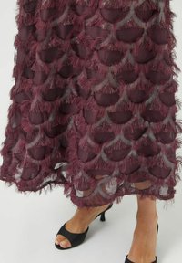 Burgunderfarvet kjole med lagdelt, tekstureret frynse i bølgemønster. Flydende, gennemskinneligt stof. Sorte åbne sko med lav hæl synlig.