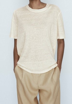 Jasno-beżowa koszulka z okrągłym dekoltem, wykonana z miękkiego, strukturalnego materiału; luźny krój z krótkimi rękawami i zaokrąglonym dołem. Połączona z beżowymi spodniami.