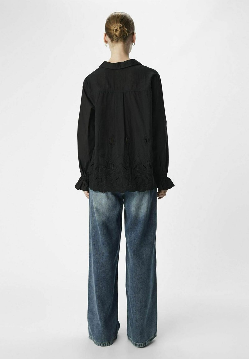 Femme avec les cheveux en chignon portant un blouse noire à manches longues avec un ourlet brodé et un jean en denim bleu délavé ample, debout de dos sur fond blanc.