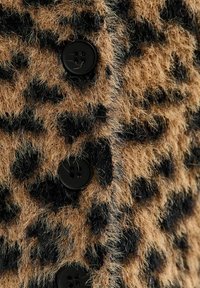 Luipaardprint zachte stof met zwarte vlekken, voorzien van zwarte knopen. De textuur lijkt zacht en pluizig, wat een gedurfd patroon creëert.