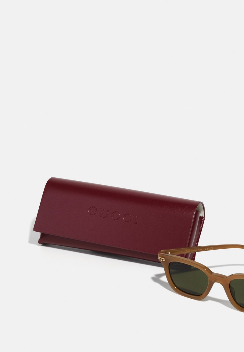 Custodia rigida bordeaux Gucci con logo in rilievo accanto a occhiali da sole marrone chiaro con lenti verdi e design angolare.