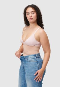 Jasnoróżowy biustonosz typu bralette z cienkimi ramiączkami w połączeniu z jasnoniebieskimi, przetartymi dżinsami trzymanymi za talię, pokazującymi luźny krój.