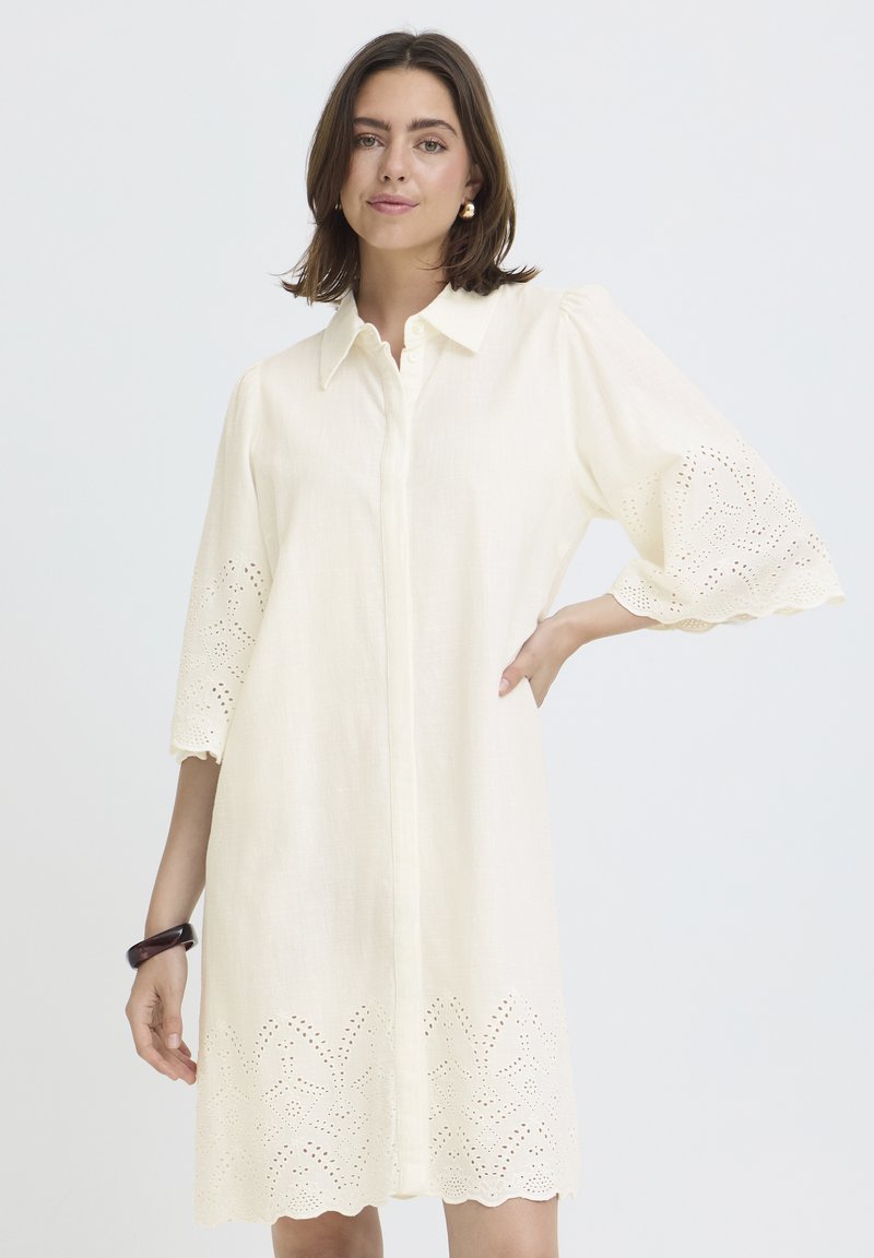 Femme portant une robe chemise crème avec des manches 3/4 en broderie anglaise et un ourlet, debout avec une main sur la hanche devant un fond blanc.