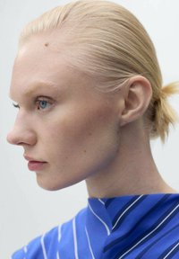 Profile view of a person with light blonde hair styled in a low ponytail, wearing a blue shirt with white vertical stripes. Smooth skin texture.