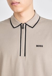 Beige polo tričko s zipovým zapínáním, černé akcenty na límci a logo na levé hrudi. Hladká textura, ležérní design.