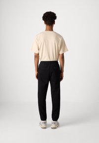 Hombre de pie de espaldas, vistiendo una camisa de manga corta color beige, pantalones jogger negros con bolsillo trasero y zapatillas blancas, sobre un fondo liso.