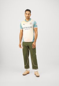 Sporttrikot in Pastellfarben mit einer hellrosa Vorderseite, blauen Ärmeln und schwarzer Schrift. Kombiniert mit olivgrünen Hosen mit crèmefarbenen Akzenten.