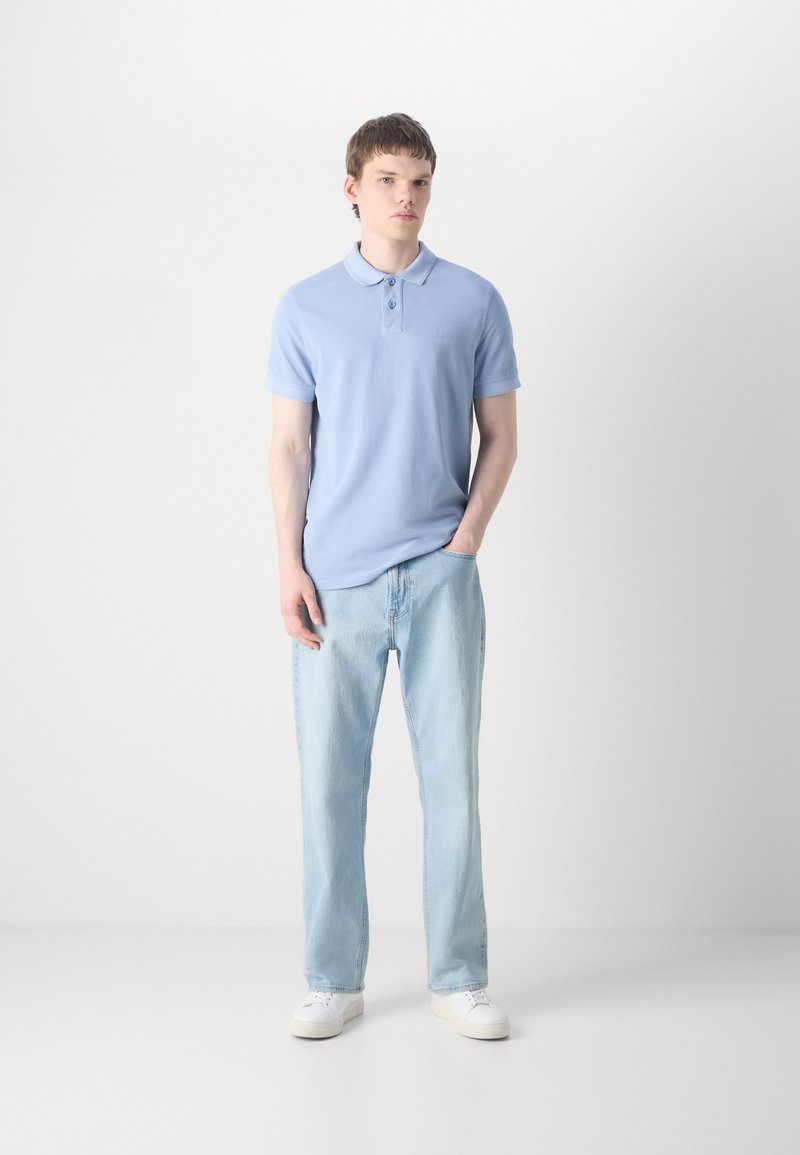 Polo shirt bleu clair avec trois boutons, associé à un jean en denim clair. Le modèle porte des baskets blanches. Design simple, texture de tissu lisse.