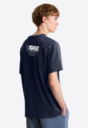 Junger Mann mit hellbraunen Haaren, der ein marineblaues T-Shirt mit dem Logo "1966 The North Face" auf dem Rücken trägt, die Hände in den Taschen, nach rechts schauend.