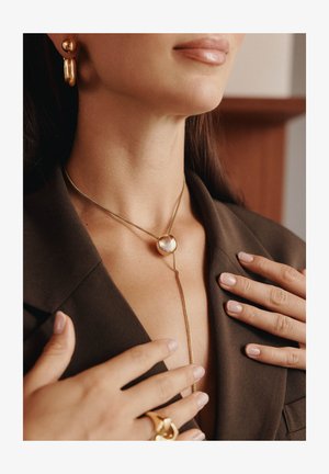 Goldene Halskette mit einem runden Anhänger, die auf einem dunklen Blazer liegt, kombiniert mit goldenen Creolen, die Hände sanft am Ausschnitt berührend.