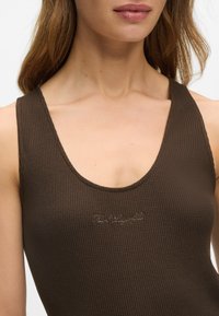 Body din material ribbed, de culoare caramel, cu bretele subțiri și decolteu rotund, având un detaliu brodat cu logo-ul în centru. Textură de țesătură netedă.