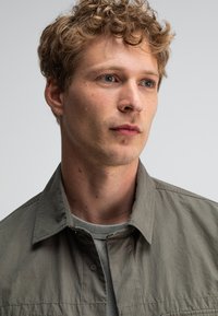 Lichtgrijze coll jacket met een gestructureerde, matte afwerking, gedragen over een lichtgroen shirt. Het onderwerp heeft golvend, kort haar en een neutrale uitstraling.