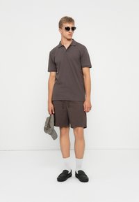 Ung man i mörkbrun pikétröja och shorts, som håller en grå keps, bär svarta skor, vita strumpor och solglasögon, står mot en vit vägg.