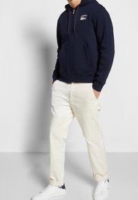 Námořnická zipová mikina s klokankovou kapucí a bílým logem; doplněná krémovými kalhotami a bílými teniskami s námořnickým detailem.