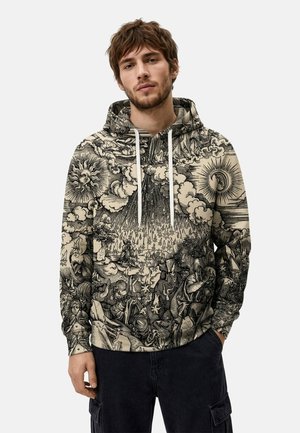 Mann trägt schwarzen und beigefarbenen Hoodie mit detailreichem Druck eines himmlischen und mythischen Szenenbildes, steht mit einer Hand in der Tasche vor einfarbigem Hintergrund.