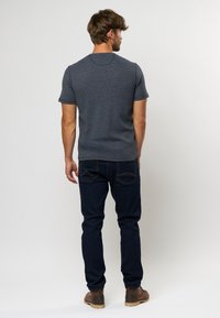 Camiseta gris de cuello redondo con mangas cortas, de tela texturizada, combinada con jeans de mezclilla oscuros y zapatos marrones, vista desde la parte de atrás.