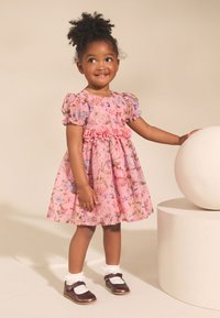 Roosa lillemustriga kleit puff-varrukate, volangilise vöökohaga ja täispika seelikuga. Sobitatud valgete pahkluu sokkide ja läikivate pruuni jalanõudega. Kõrval ringikujuline rekvisiit.