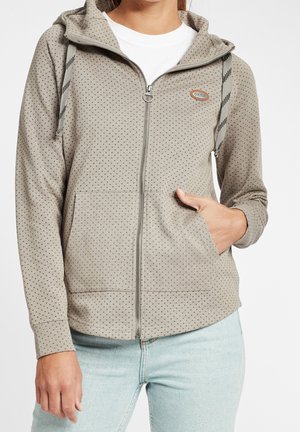 Sweat à capuche beige zippé avec petits pois noirs, cordons rayés, poche kangourou, porté sur un t-shirt blanc et un jean bleu clair.