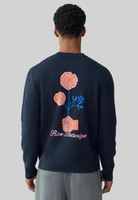 Pulover tricotat bleumarin cu un design floral pe spate, cu flori roz, tulpini albastre și o ilustrație a unei mâini. Textul spune „Rêve Botanique”.