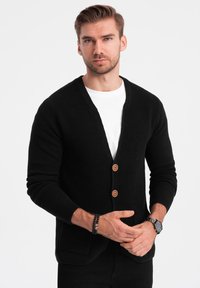 Cárdigan negro de punto con cuello en V, mangas largas, dos botones y bolsillos delanteros, usado sobre una camiseta blanca. Textura y estructura sutiles.