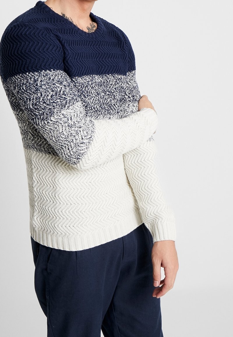 Gebreide trui met een zigzag patroon, met een marineblauw bovenstuk, witte middelste sectie en cremekleurige onderkant. Ribgebreide manchetten en onderrand voegen textuur toe.