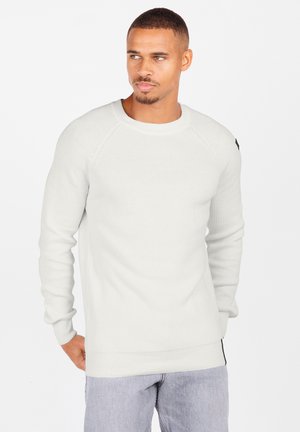 Ribbed white sweater with long sleeves, round neck, and black accent on left shoulder. Fabric appears soft and slightly textured.