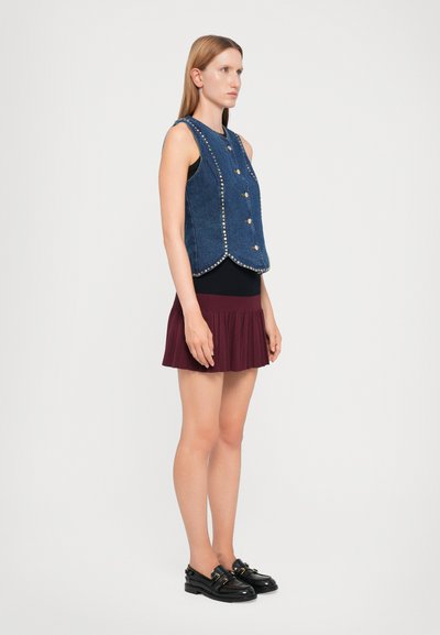 Denim veste ar sudraba kniedēm, pieguļošs dizains, seši pogas, valkāta virs melnas blūzes, kombinēta ar bordo plisētu svārku un melnām kurpēm.