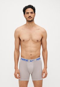 Sivi Nike bokserici s plavim elastičnim pojasom, koji imaju uski kroj i glatku teksturu tkanine. Nema uzoraka niti dodatnog hardvera vidljivog.