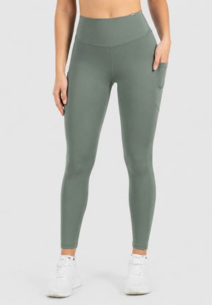 Femme portant des leggings taille haute vert olive avec une poche latérale et des baskets blanches, debout devant un fond uni.