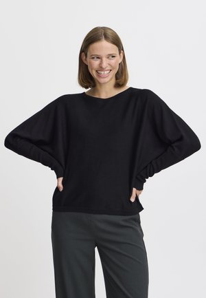 Un pull noir surdimensionné avec des manches larges et des poignets côtelés, présentant une coupe décontractée et une texture lisse. Associé à un pantalon sombre.