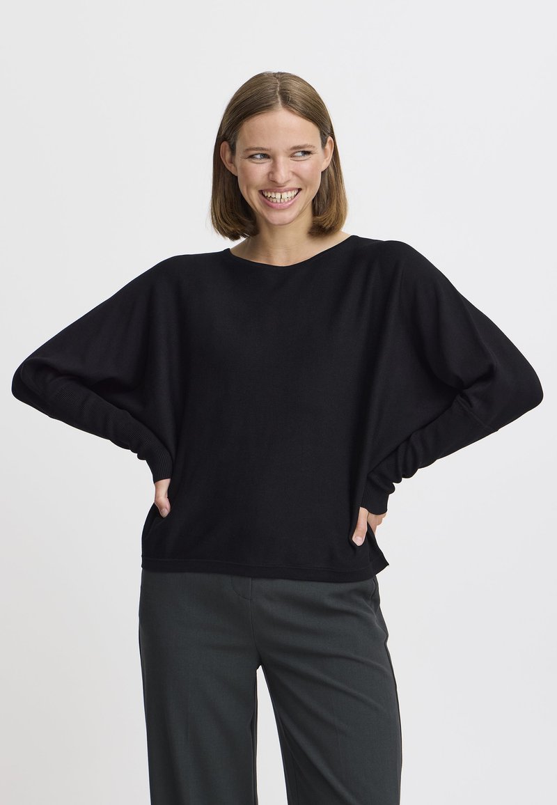 Un pull noir surdimensionné avec des manches larges et des poignets côtelés, présentant une coupe décontractée et une texture lisse. Associé à un pantalon sombre.