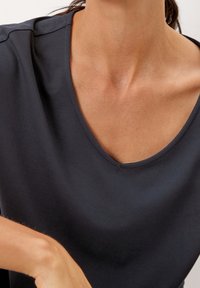 Frau in einem dunkelblauen V-Ausschnittshirt, das den Ausschnitt, die Schlüsselbeine und einen Teil ihres Arms vor einem hellen Hintergrund zeigt.